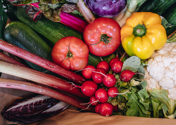 Eine mit Papier ausgeschlagene Gemüsekiste, die mit Gurke, Blumenkohl, Paprika, Radieschen, Lauchzwiebeln, Kohlrabi und Tomaten befüllt ist.nd Radieschen gefüllt ist. Eine Mitarbeiterin legt noch einen blauen Kohlrabi dazu.