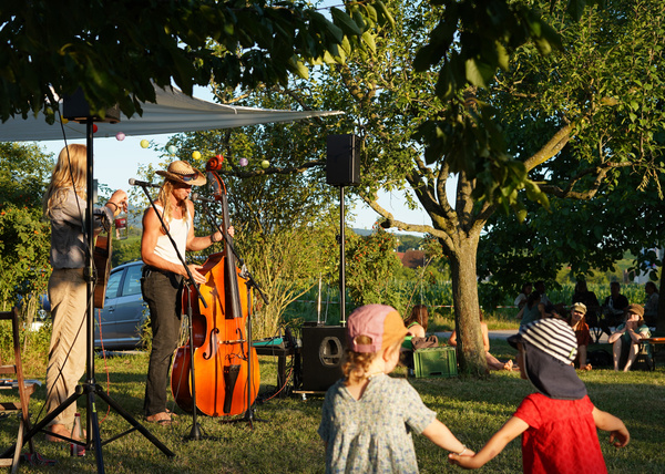 KI generiert: Konzert im Freien: Musiker spielen unter Bäumen, während Kinder im Vordergrund tanzen.