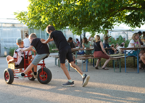 KI generiert: Kinder spielen auf einem dreirädrigen Gefährt, während Erwachsene an Tischen im Freien sitzen.