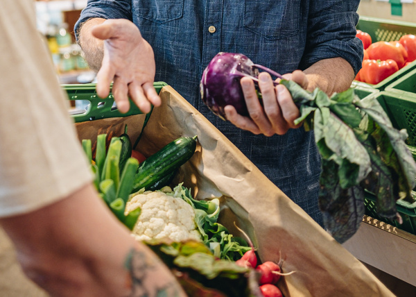 Eine mit Papier ausgeschlagene Gemüsekiste, die mit Gurke, Blumenkohl und Radieschen gefüllt ist. Eine Mitarbeiterin legt noch einen blauen Kohlrabi dazu.