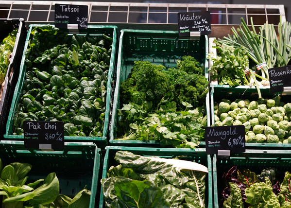 KI generiert: Verschiedene Gemüsesorten in Kisten auf Markt; Schilder mit Preisen: Tatsoi, Pak Choi, Mangold, Kräuter, Rosenkohl.