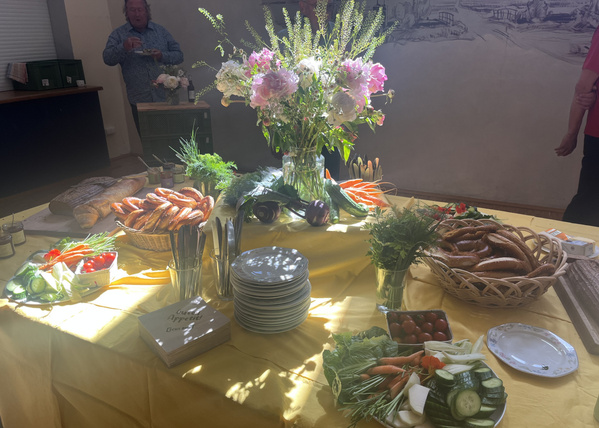 KI generiert: Buffet mit Blumenstrauß, Brot, Brezeln, Gemüse und Geschirr auf einem gelben Tisch. Text: „Guten Appetit“.