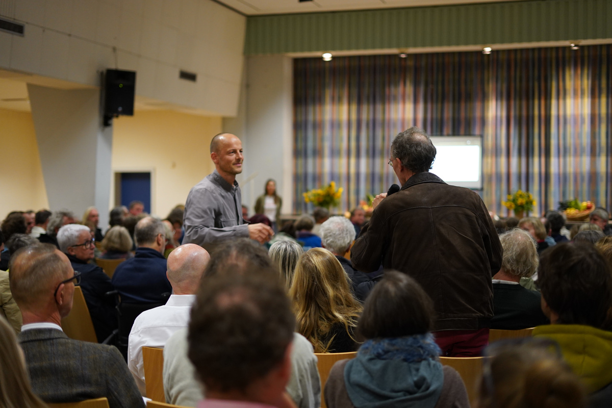 KI generiert: Ein Mann spricht in einem voll besetzten Saal ins Mikrofon, während ein anderer Mann zuhört.