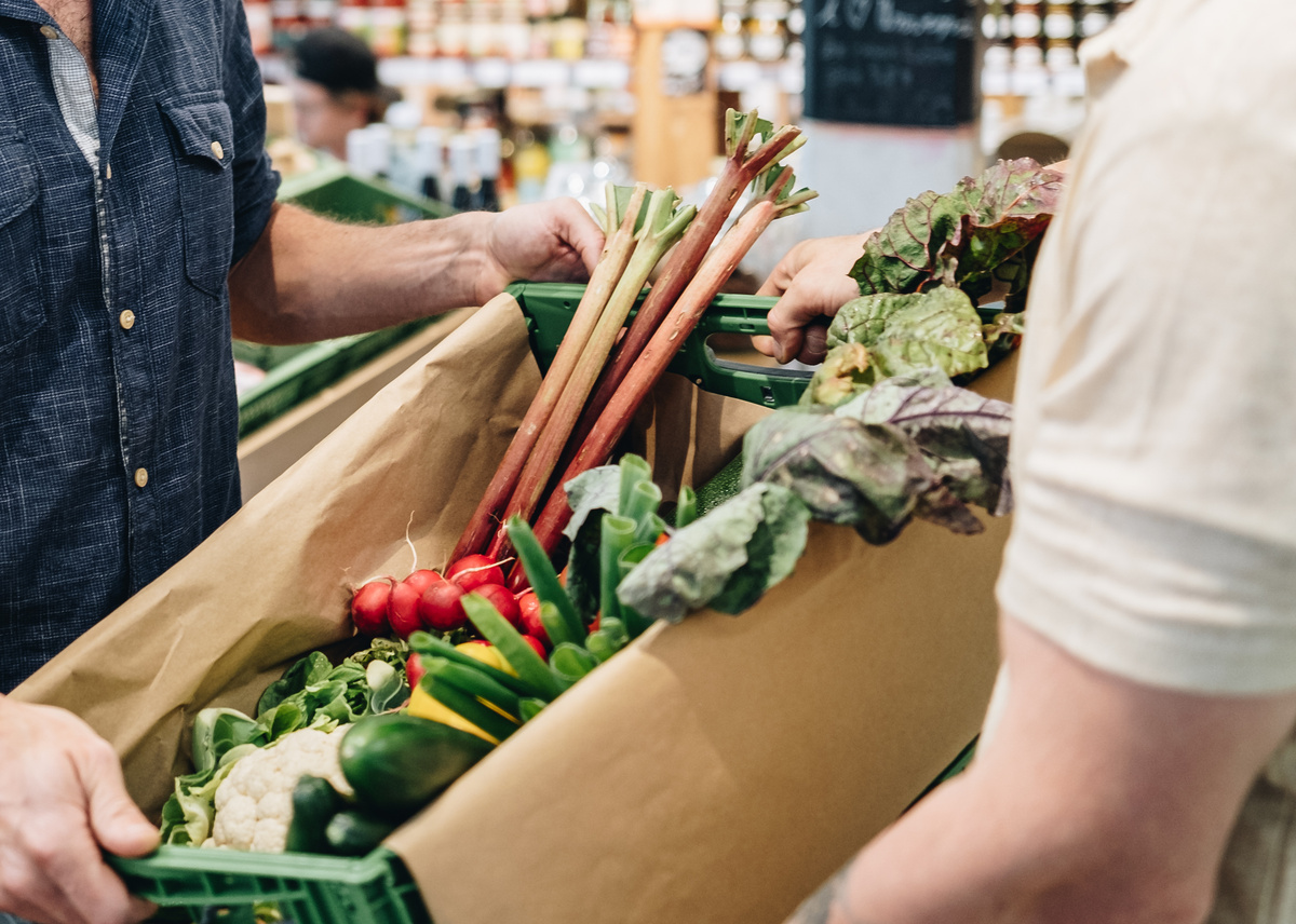 KI generiert: Eine Gemüsekiste voller frischer Produkte in einem Laden, zwei Personen tragen sie gemeinsam.
