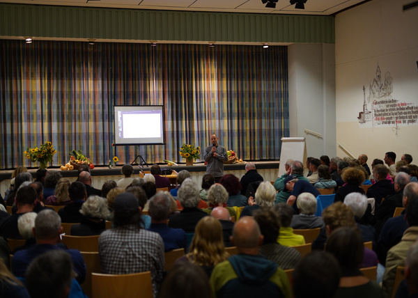 KI generiert: Ein Vortragender spricht vor einem Publikum in einem Saal. Links ist eine Projektionsfläche sichtbar.