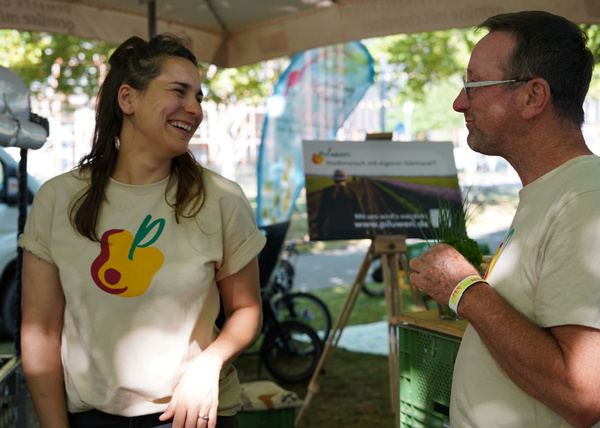 KI generiert: Zwei Personen unterhalten sich an einem Stand im Freien. Im Hintergrund ist ein Plakat zu sehen.